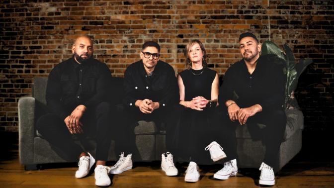 The 4 band members of dehli 2 dublin seated on a couch in front wearing all black with white sneakers