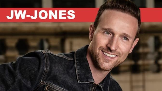 smiling musician JW Jones wearing a black jean jacket and blue jeans holding his guitar