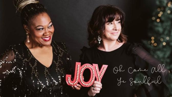 Vocalist Dawn Pemberton smiling in a sparkling black holiday dress holding a red sign that says Joy, standing beside Bassist Jodi Proznick smiling and wearing a black holiday dress beside a christmas tree