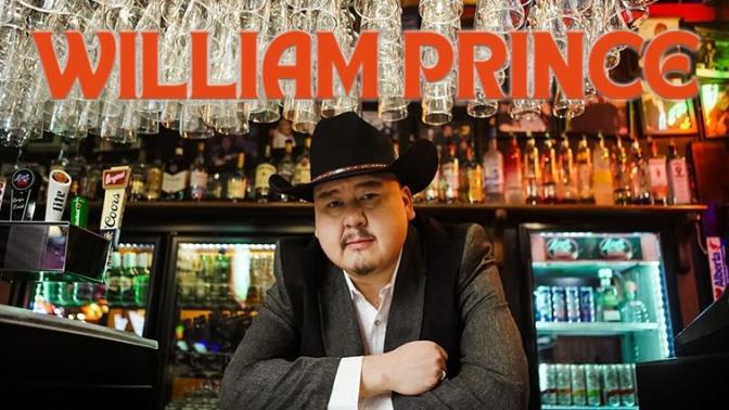 William Prince seated at a bar in a western suit and hat, with glasses hanging above and bottles in the background