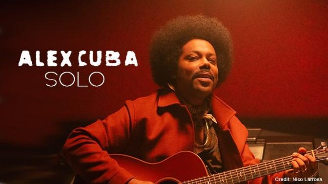 Latin Pop artist Alex Cuba seated with guitar in a burnt orange suit and room