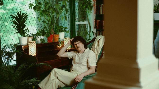 Boy Golden, Musical persona of LIam Duncan seated in a retro styled room with plants and green tile, weaing a beige leisure coordinated pant set