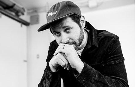 Black and white photo of a man wearing a baseball cap and jacket, resting his chin on his hands and looking thoughtfully into the camera.