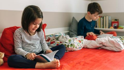 two children sit under blankets in their pijamas reading their books.
