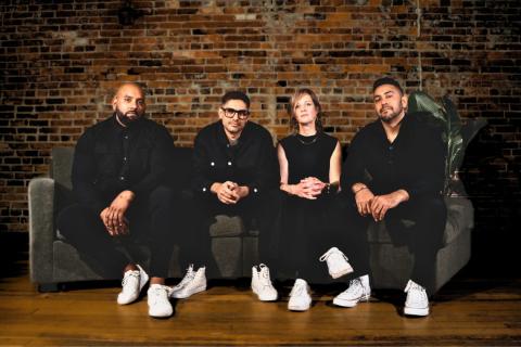 The 4 band members of dehli 2 dublin seated on a couch in front wearing all black with white sneakers