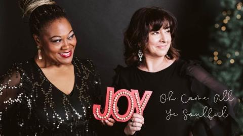 Vocalist Dawn Pemberton smiling in a sparkling black holiday dress holding a red sign that says Joy, standing beside Bassist Jodi Proznick smiling and wearing a black holiday dress beside a christmas tree