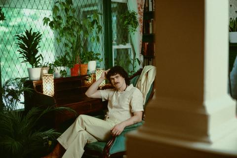 Boy Golden, Musical persona of LIam Duncan seated in a retro styled room with plants and green tile, weaing a beige leisure coordinated pant set