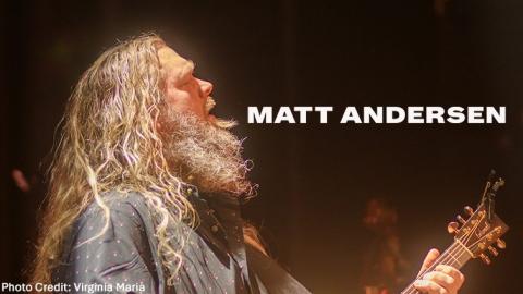 Musician Matt Andersen in profile while singing and playing guitar on stage. Matt has long wavy salt and pepper hair and a beard